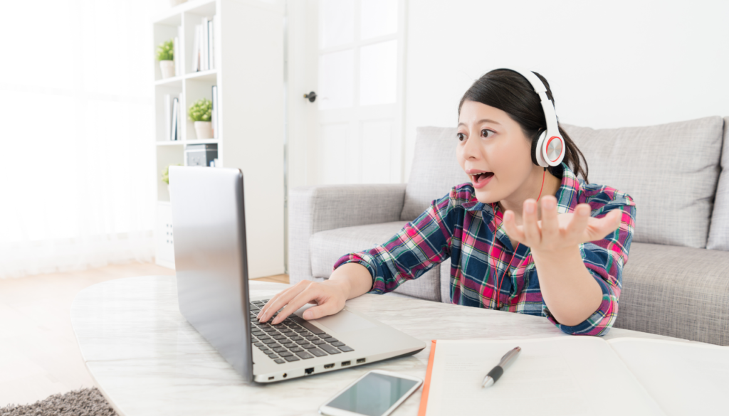 Jovem usando fones de ouvido em uma videochamada, interagindo com entusiasmo em frente a um laptop, com livros e anotações sobre a mesa.