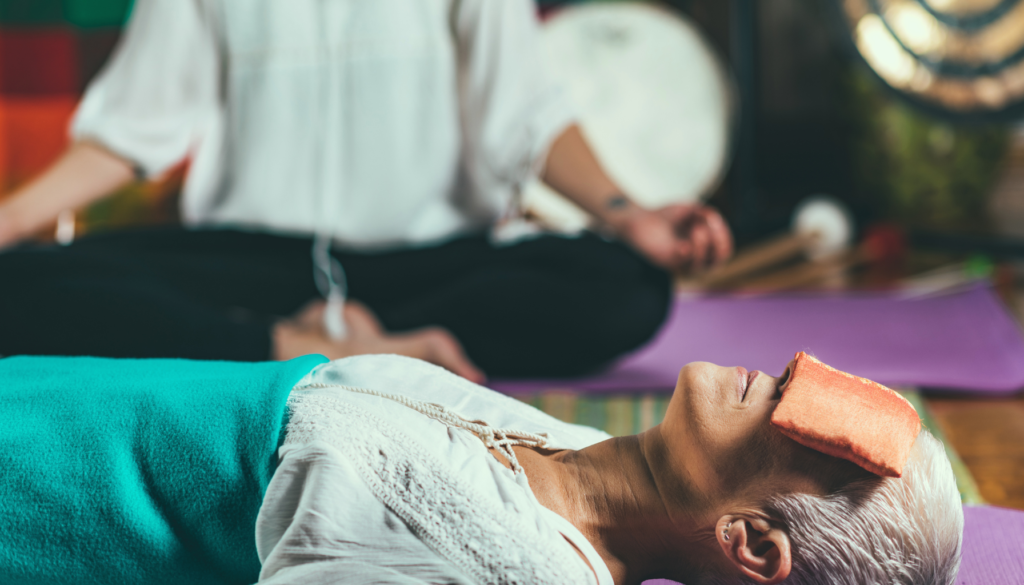 Pessoa idosa relaxando em sessão de terapia energética, coberta com manta azul e pano laranja sobre os olhos, enquanto terapeuta medita ao fundo em um ambiente acolhedor.