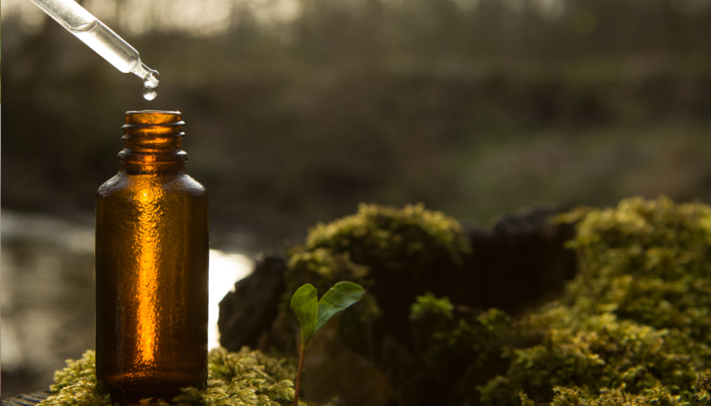 Frasco de vidro âmbar com gotas de óleo essencial caindo de um conta-gotas, em um ambiente natural com musgos e plantas ao fundo.