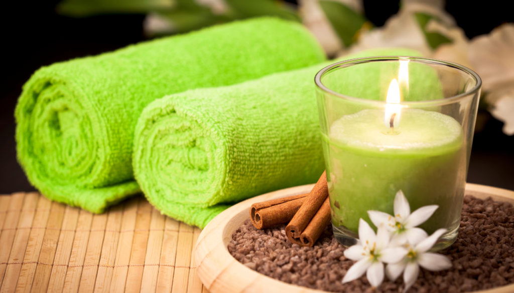 Ambiente Relaxante com Aromaterapia Toalhas verdes, vela aromática acesa, canela e flores brancas sobre uma bandeja de bambu, compondo um ambiente relaxante.