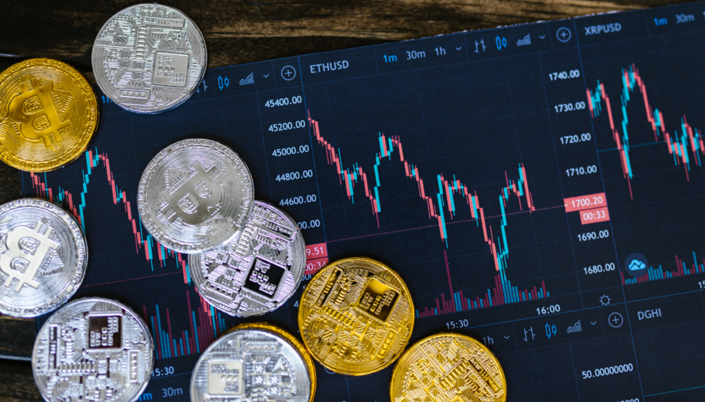Moedas físicas representando Bitcoin em prata e ouro dispostas sobre uma tela mostrando gráficos de preços de criptomoedas em um ambiente de negociação.