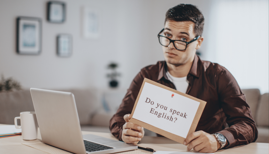 Como Construir um Vocabulário Sólido em Inglês Homem sentado em frente a um laptop segurando um cartaz com a frase "Do you speak English?", representando a importância de aprender como é que se fala inglês.