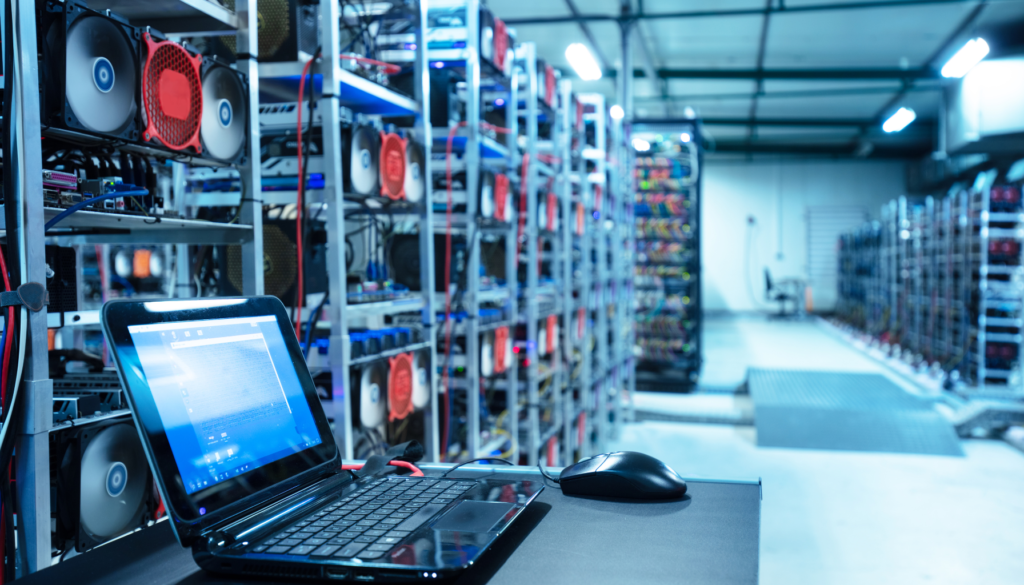 Mineração de Criptomoedas: A Infraestrutura por Trás da Blockchain Um data center de mineração de criptomoedas, com racks de GPUs e sistemas de refrigeração, acompanhado por um laptop aberto para monitoramento.