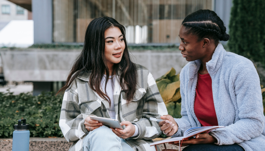 Aprenda Inglês com Amizade e Prática Duas mulheres sentadas ao ar livre, conversando e estudando juntas, com uma segurando um tablet e a outra um caderno.