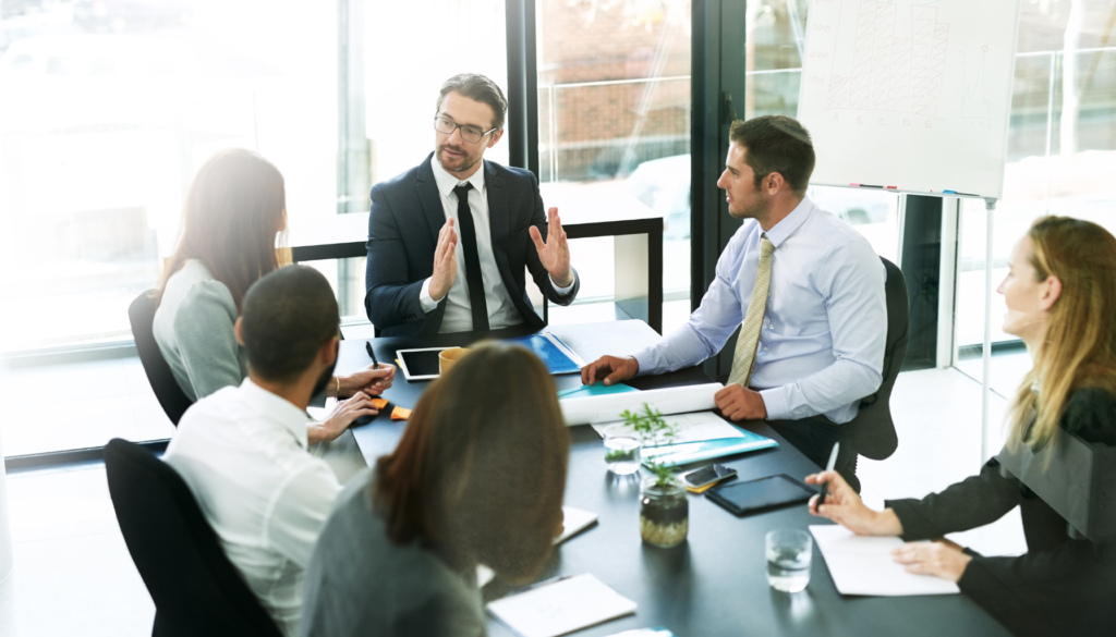 Comunicação Eficaz: A Chave para Reuniões Produtivas Grupo de profissionais em uma reunião, com um homem gesticulando ao centro, ilustrando a importância da comunicação eficaz em ambientes corporativos.