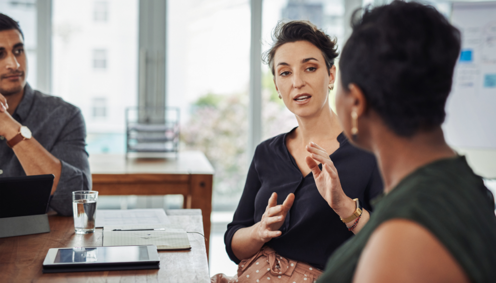 Comunicação Eficaz: Transforme Diálogos em Soluções Mulher explicando ideias durante uma reunião em um ambiente de escritório, enquanto outra pessoa ouve atentamente.