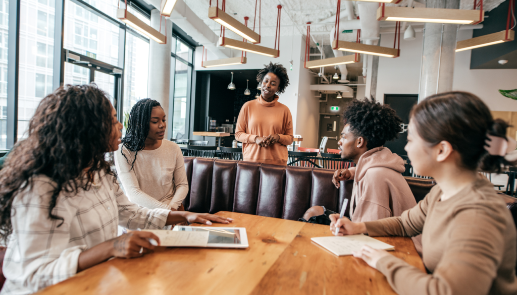 Comunicação Eficaz: Engajando Equipes e Inspirando Resultados Grupo de pessoas em um ambiente descontraído, participando de uma reunião colaborativa enquanto uma mulher apresenta suas ideias.
