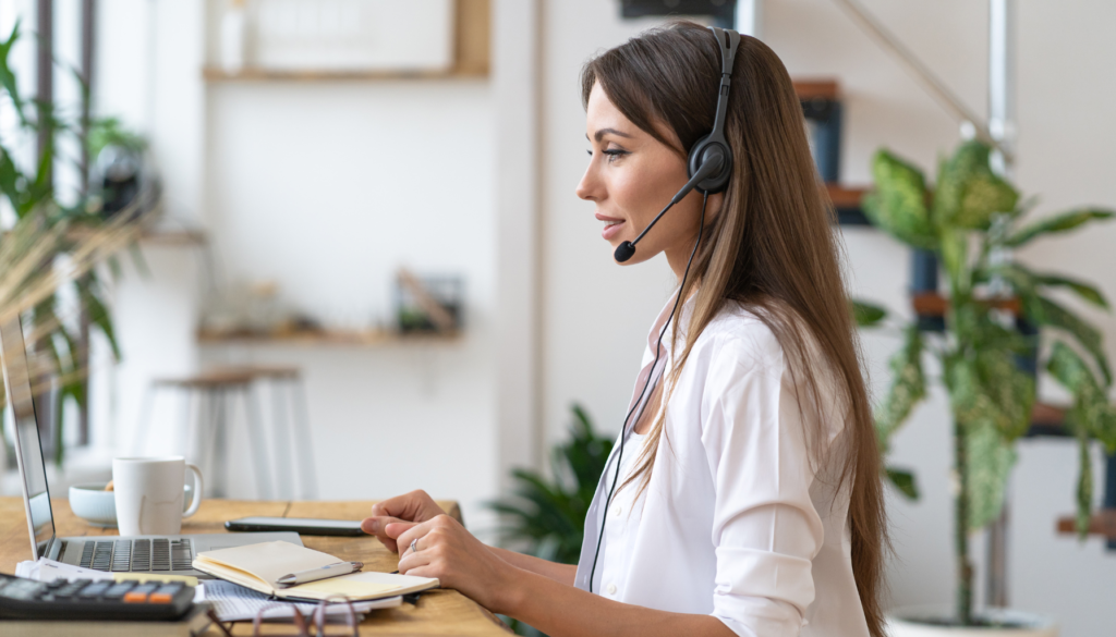 Mulher usando headset, trabalhando em um ambiente de escritório moderno com laptop, papéis e plantas ao fundo, representando comunicação eficiente em atendimento.