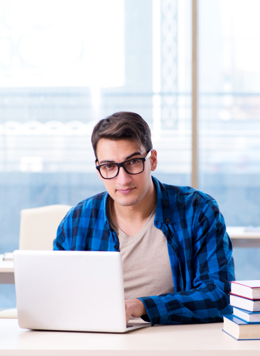 Jovem estudando com laptop e livros empilhados em uma mesa, em um ambiente moderno e iluminado.

