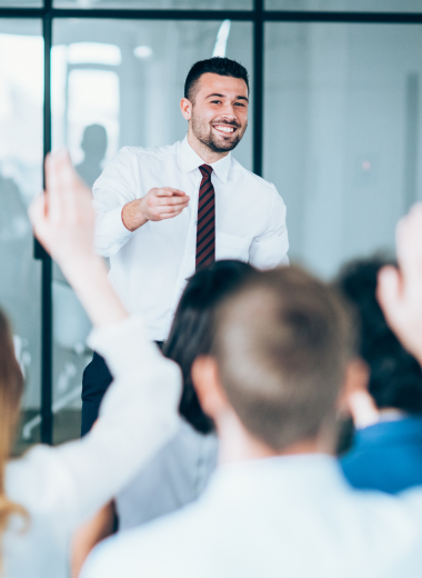 Palestrante sorridente interagindo com a audiência em uma sala de conferência, com participantes levantando as mãos.

