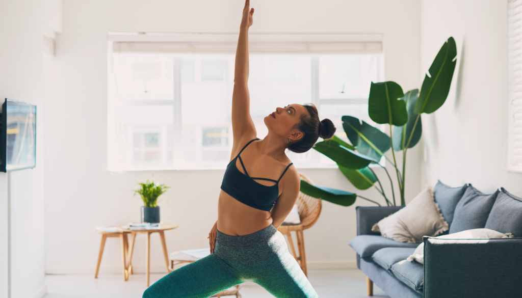 Mulher praticando yoga em uma sala iluminada com decoração minimalista, realizando uma pose com o braço estendido para cima.