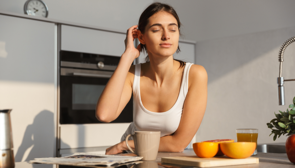 Mulher jovem em uma cozinha iluminada pelo sol da manhã, com os olhos fechados em uma expressão de tranquilidade, ao lado de frutas e uma xícara de chá.