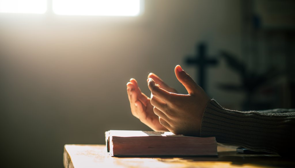 Mãos em posição de oração sobre uma Bíblia aberta, iluminadas por uma luz suave vinda de uma janela, com uma cruz desfocada ao fundo.