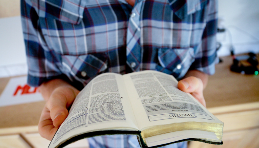 Leitura da Bíblia em 2 Timóteo Pessoa segurando uma Bíblia aberta na passagem de 2 Timóteo, vestida com camisa xadrez azul em um ambiente doméstico.
