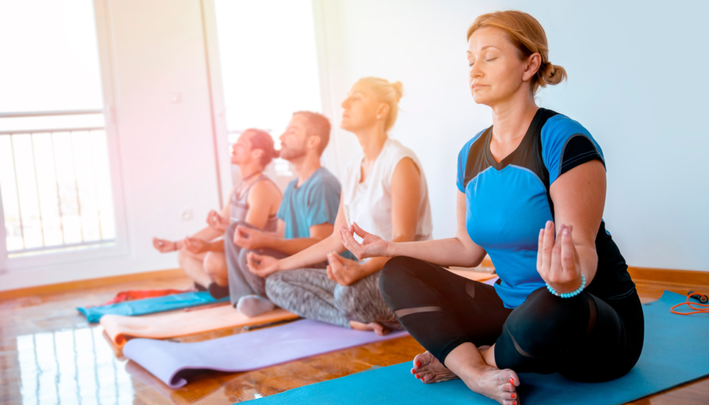 Grupo de pessoas meditando em uma sala iluminada, sentadas em tapetes de yoga, com olhos fechados e postura relaxada.