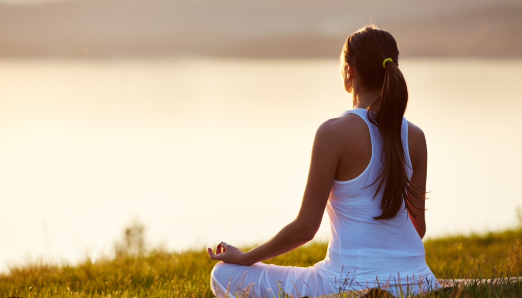 Mulher jovem sentada na grama em posição de meditação, de costas para a câmera, olhando para o horizonte ao entardecer, vestindo roupas brancas.