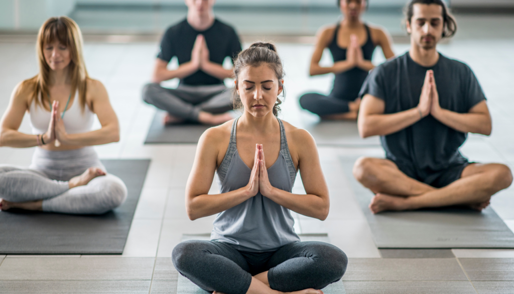 Grupo de pessoas praticando meditação em um estúdio de yoga, com uma mulher ao centro de olhos fechados e mãos unidas em posição de prece.