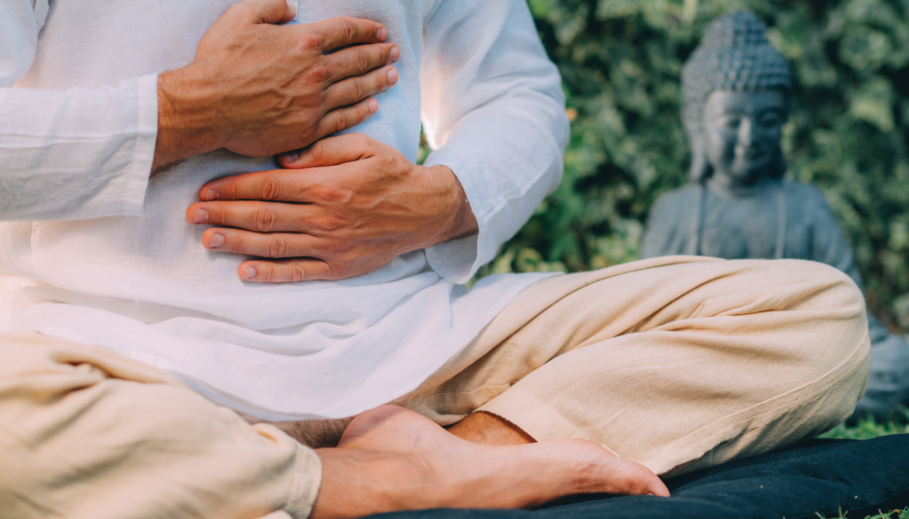 Homem sentado em posição de meditação ao ar livre, com as mãos sobre o abdômen, simbolizando conexão e cura interior.