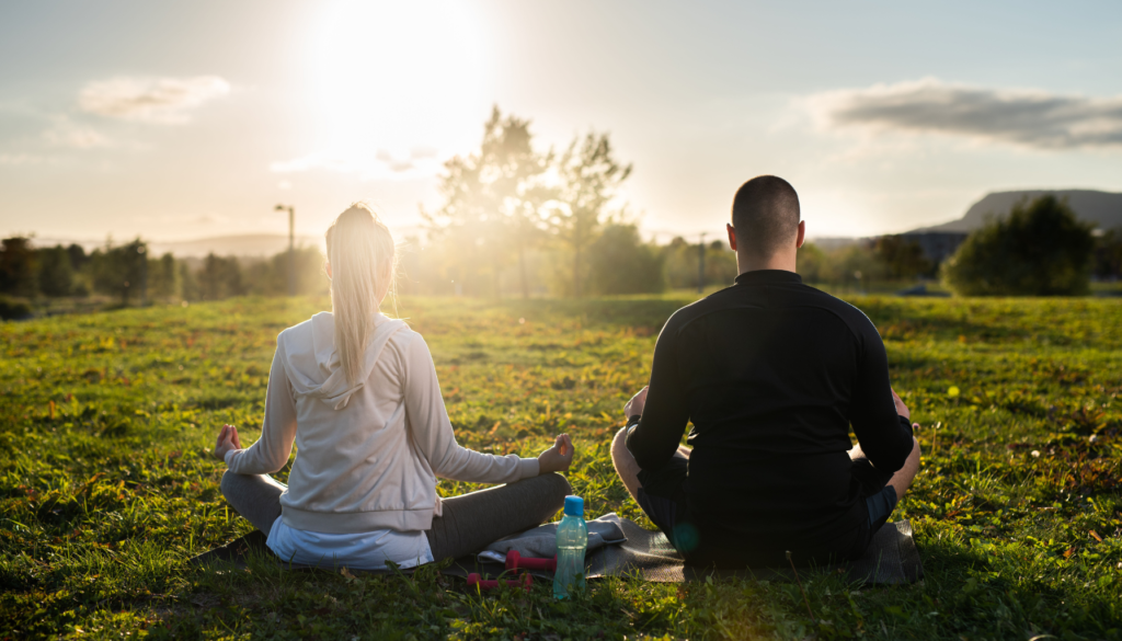 Duas pessoas meditando ao ar livre em um campo verde, sentadas de costas para a câmera, com o sol se pondo ao fundo.